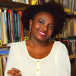Mulher negra de pele retinta, com cabelos cacheados pretos de tamanho médio, sorrindo, veste uma camisa branca de mangas compridas, ao fundo uma estante de livros.